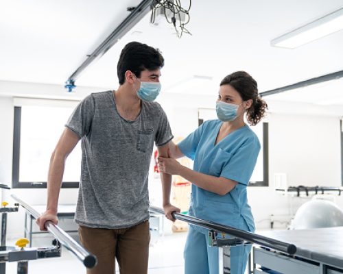 Male patient at physical rehab walking with the help of parallel bars while therapist supports him both wearing protective face masks - Pandemic lifestyles