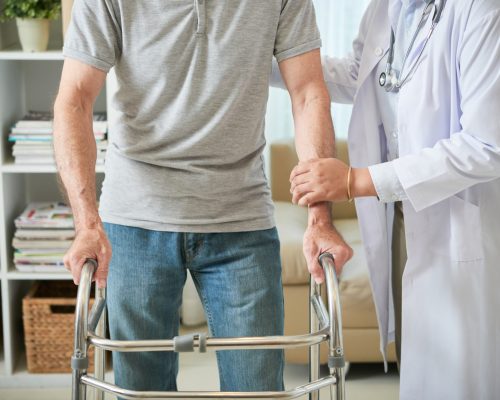 unrecognizable-female-doctor-helping-male-patient-walk-with-walking-frame